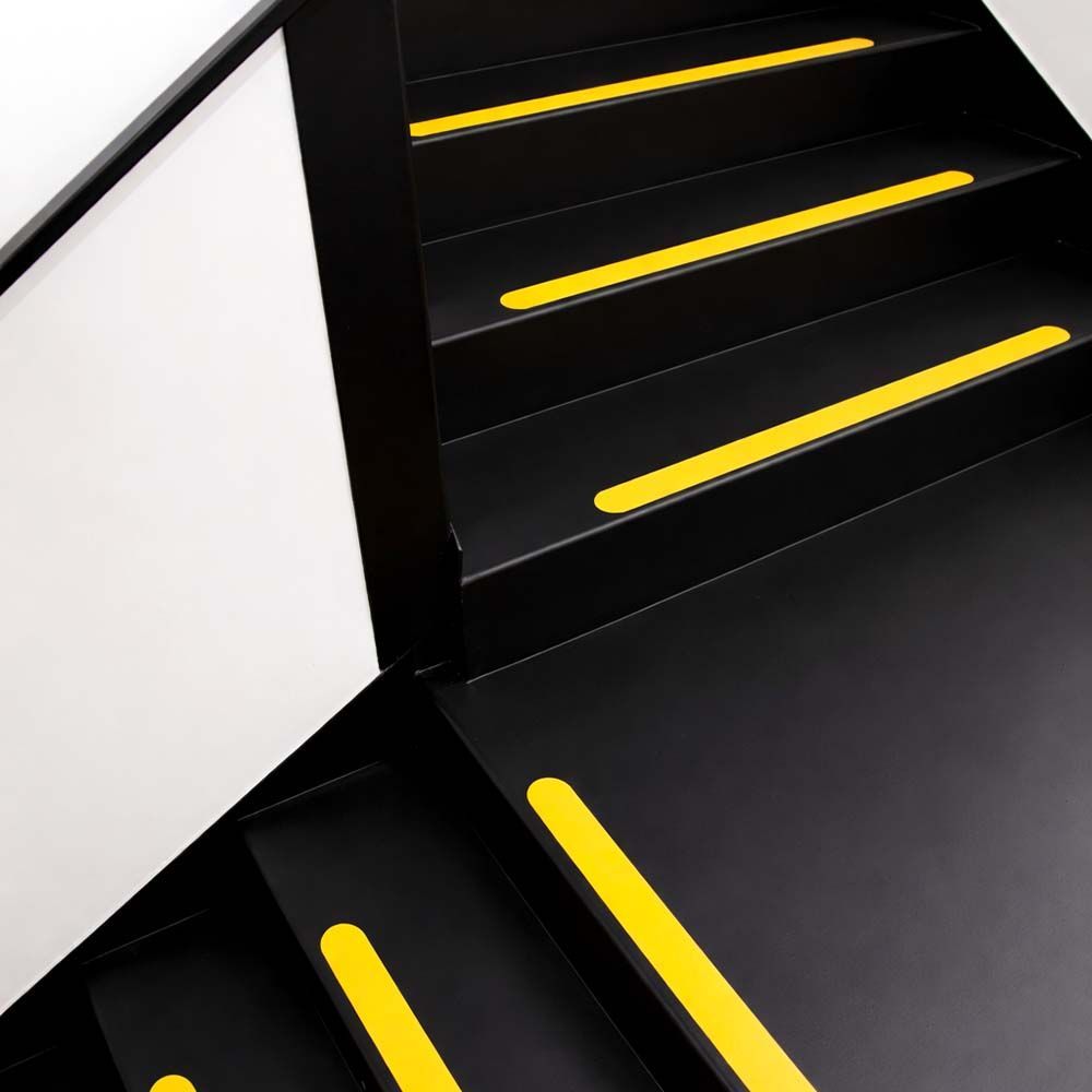 Black staircase with yellow anti-slip strips on each step, viewed from above, with white wall and handrail.