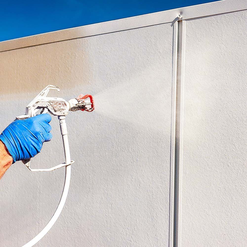 Hand in blue glove sprays paint on a textured white wall using a paint sprayer.