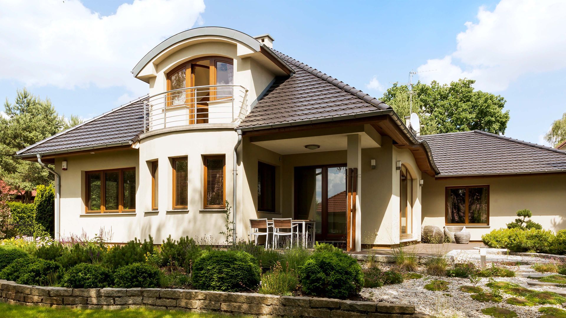 Beige house with a curved upper level, balcony, and covered porch; landscaped yard with greenery.
