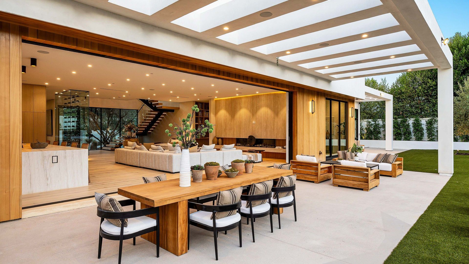 Outdoor patio with dining table, seating, and open doors to a modern living area. Sunny day, beige and brown tones.