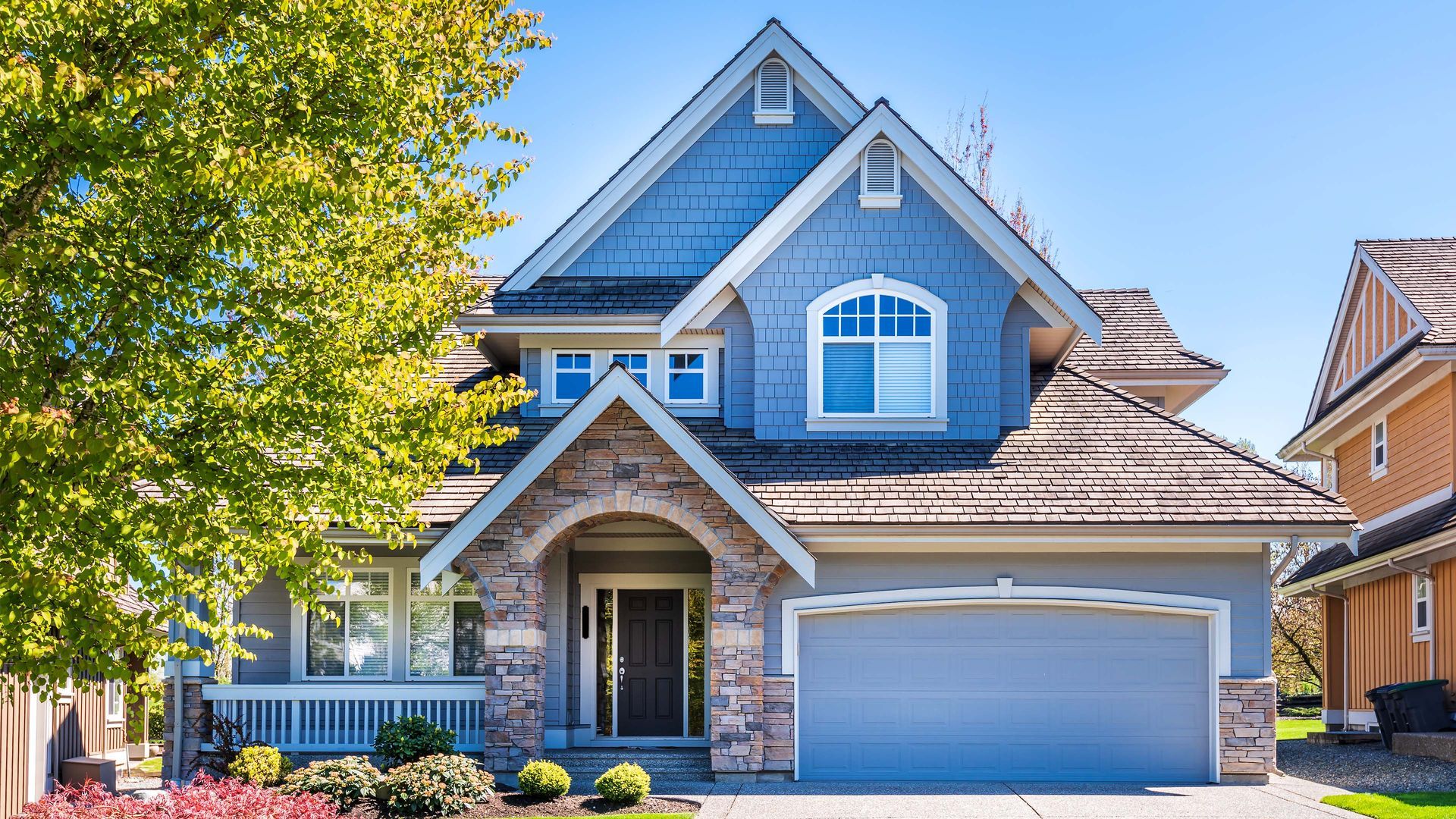 Blue house with stone accents, garage, and a lush green tree in front.