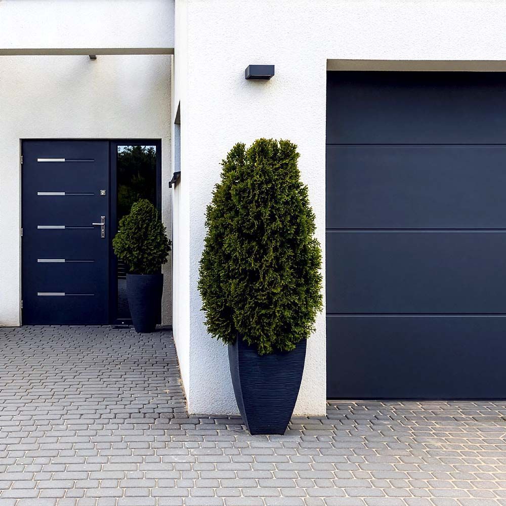 Modern home exterior with dark gray door and garage, potted evergreen trees, and gray brick walkway.