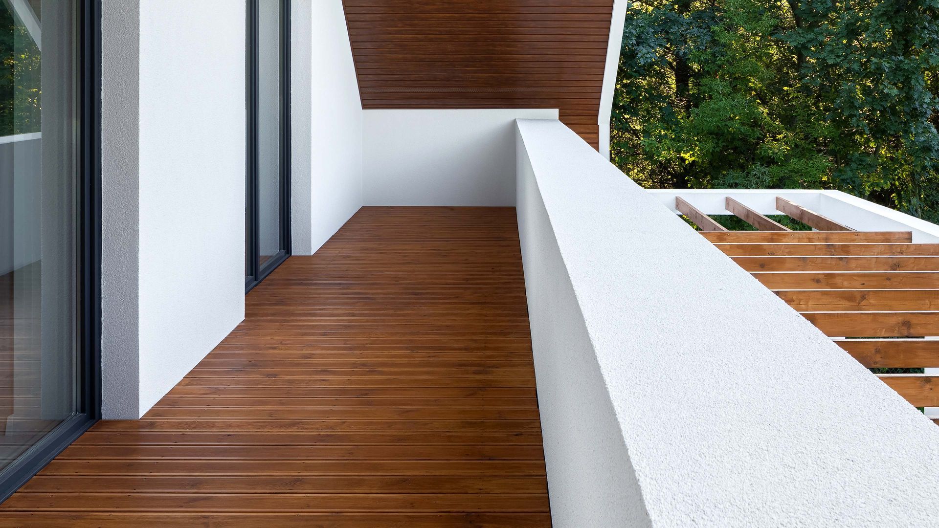 Wood-floored balcony with white walls and railing, overlooking green trees.
