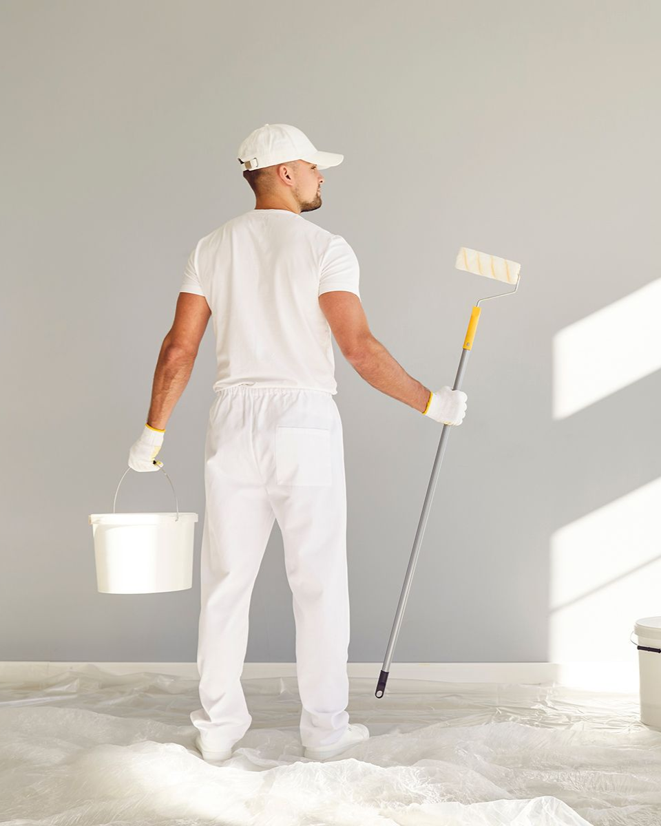 Painter in white overalls looks at wall, holding a paint roller and bucket.