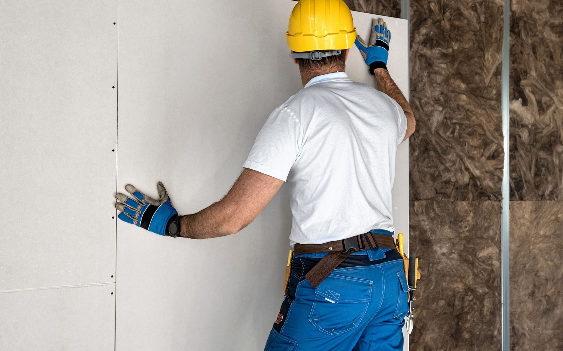 Construction worker installing drywall, wearing blue work gloves, a yellow helmet, and blue overalls.