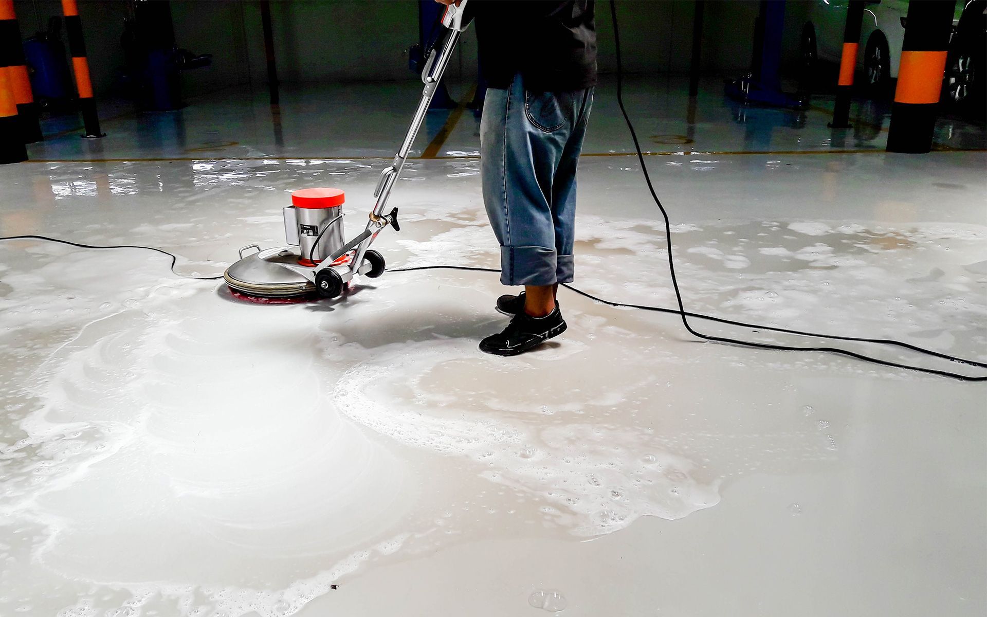 Person using a floor cleaning machine on a wet, light gray floor. Dark clothing and shoes.