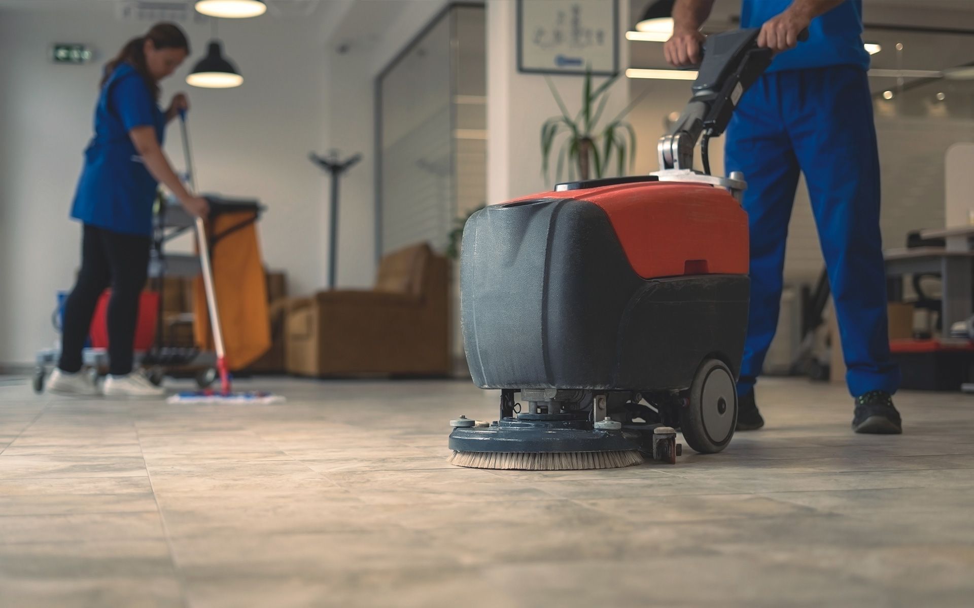 Two cleaning workers in blue uniforms: one mopping, the other operating a floor scrubber in an office.