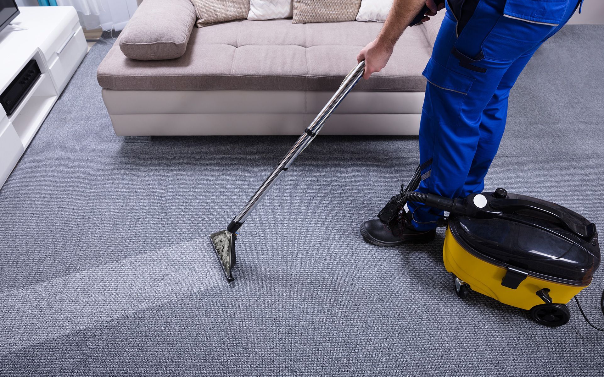 Person in blue cleaning carpet with a yellow vacuum cleaner, next to a beige couch.