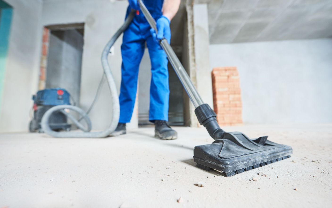 Person in blue overalls vacuuming a concrete floor in a construction site.