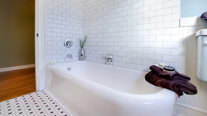 White bathtub in a tiled bathroom with a decorative rug, towels, and a vase of plants.