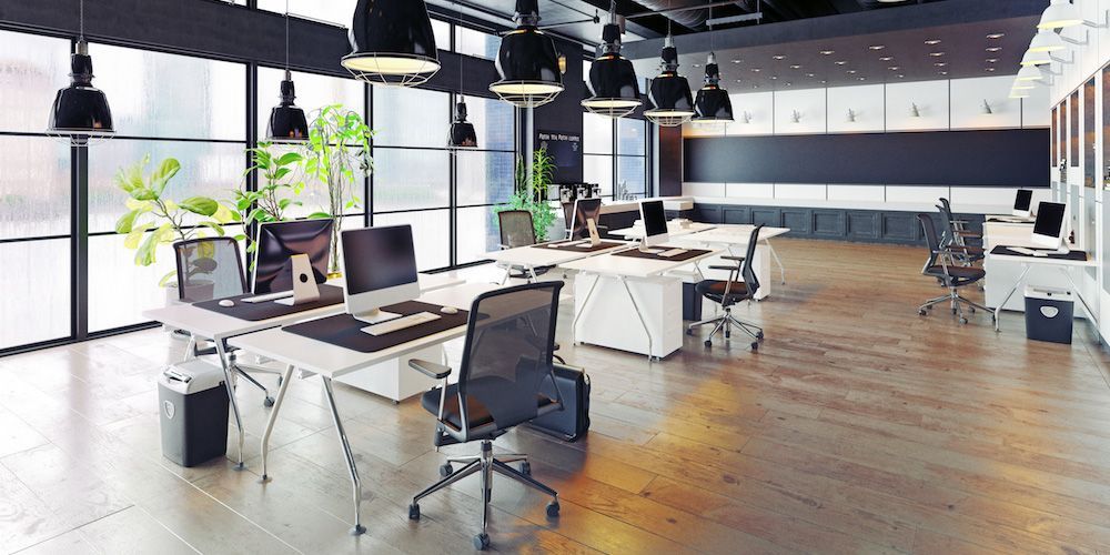Modern office space with desks, computers, chairs, and plants near large windows.