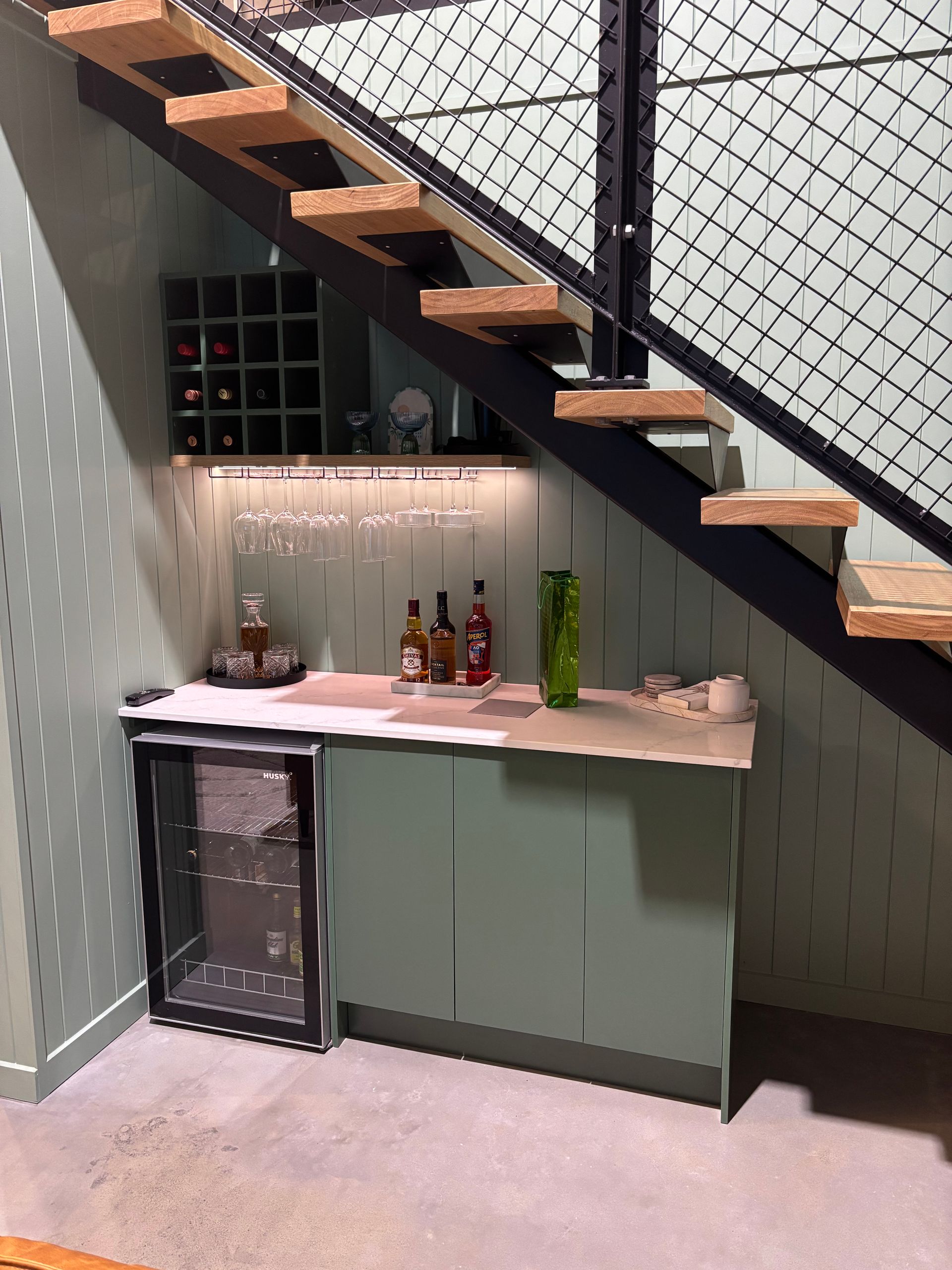 Wine bar under a staircase with a wine fridge, storage, and bottles. Light green cabinets and walls — Maken Kitchens & Cabinets in Chinderah, NSW
