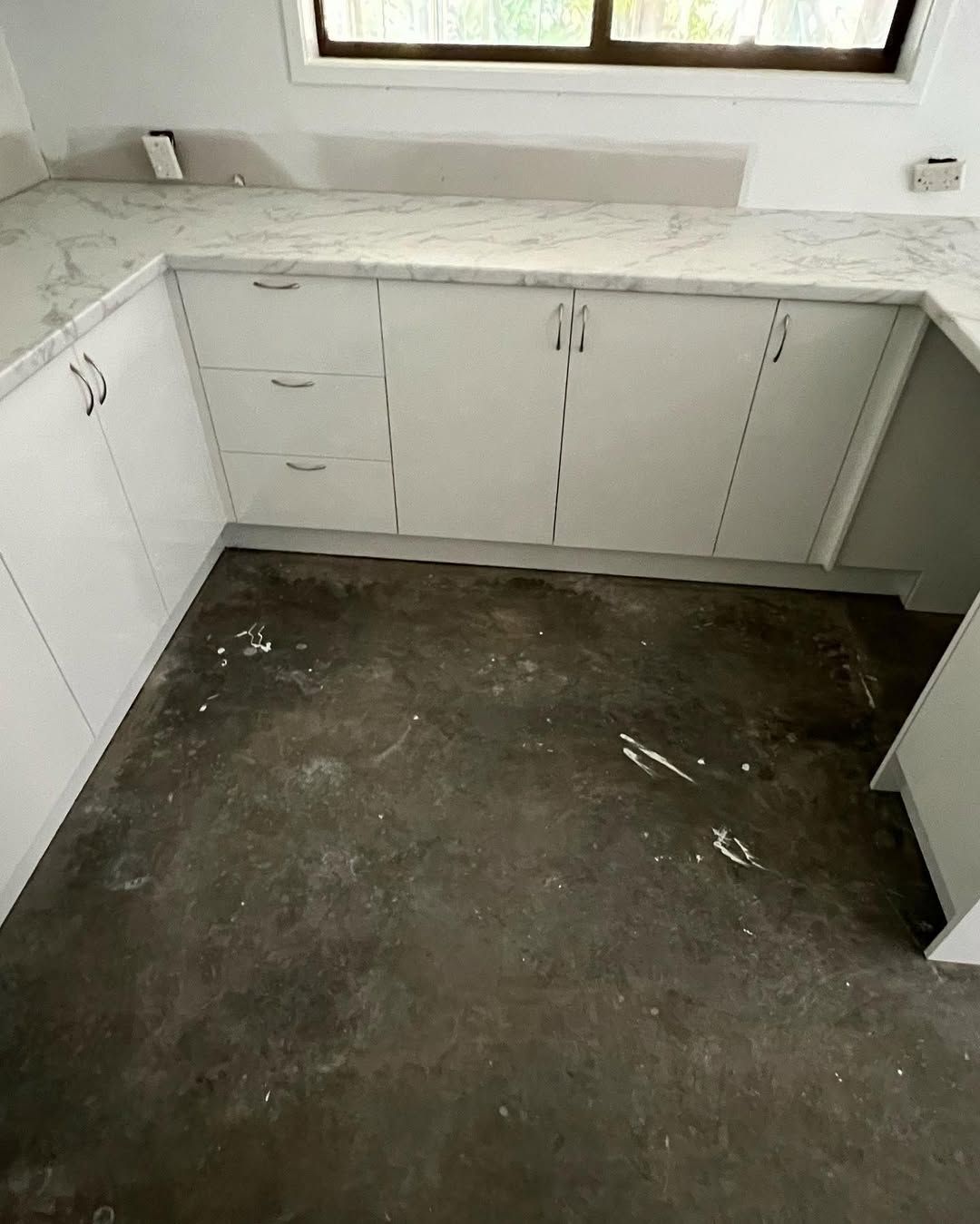 White kitchen cabinets with light countertops sit in front of a window, on a dark floor. — Maken Kitchens & Cabinets in Lismore, NSW
