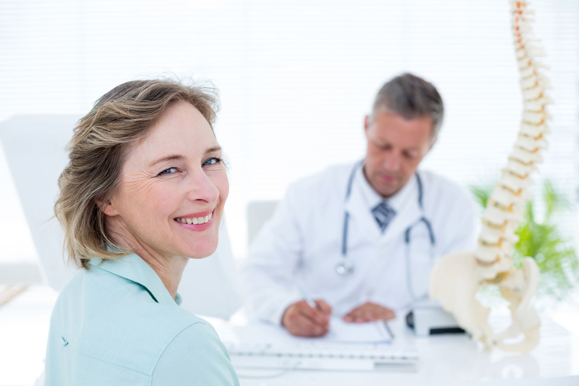 Woman with blue shirt smiles at camera with blurry doctor in background, representing urgent care. Woman with blue shirt smiles at camera with blurry doctor in background, representing urgent care.