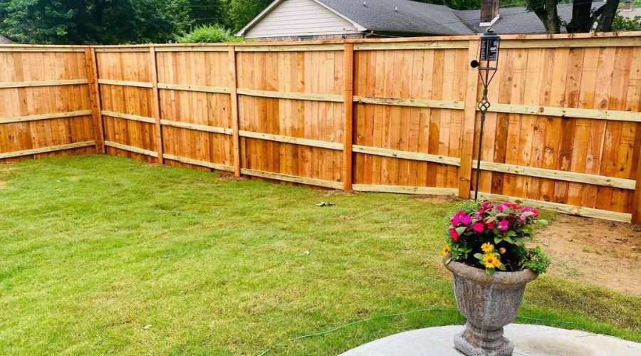 A light brown wooden privacy fence borders a grassy backyard, with a decorative stone planter in the foreground.