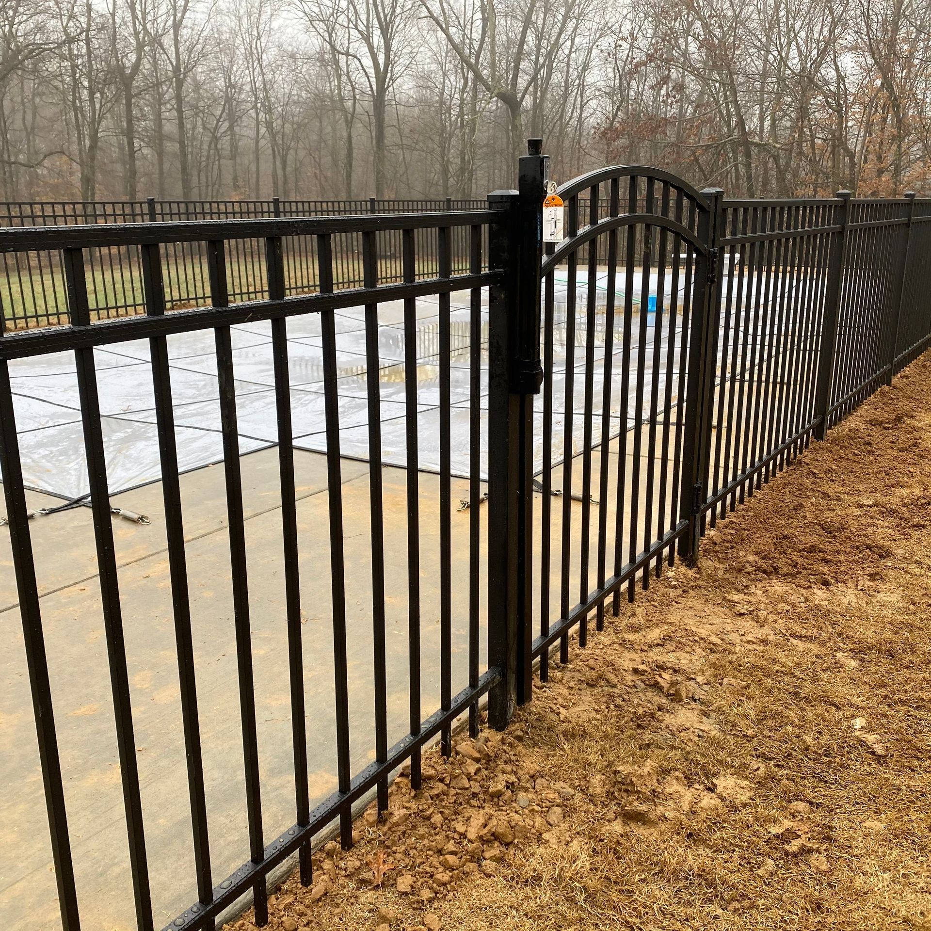 Black metal fence with gate near a pool and trees on a cloudy day.