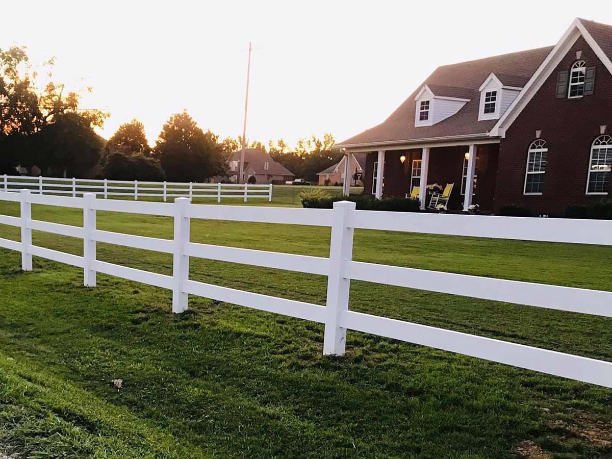 A white fence surrounds a lush green field in front of a brick house.