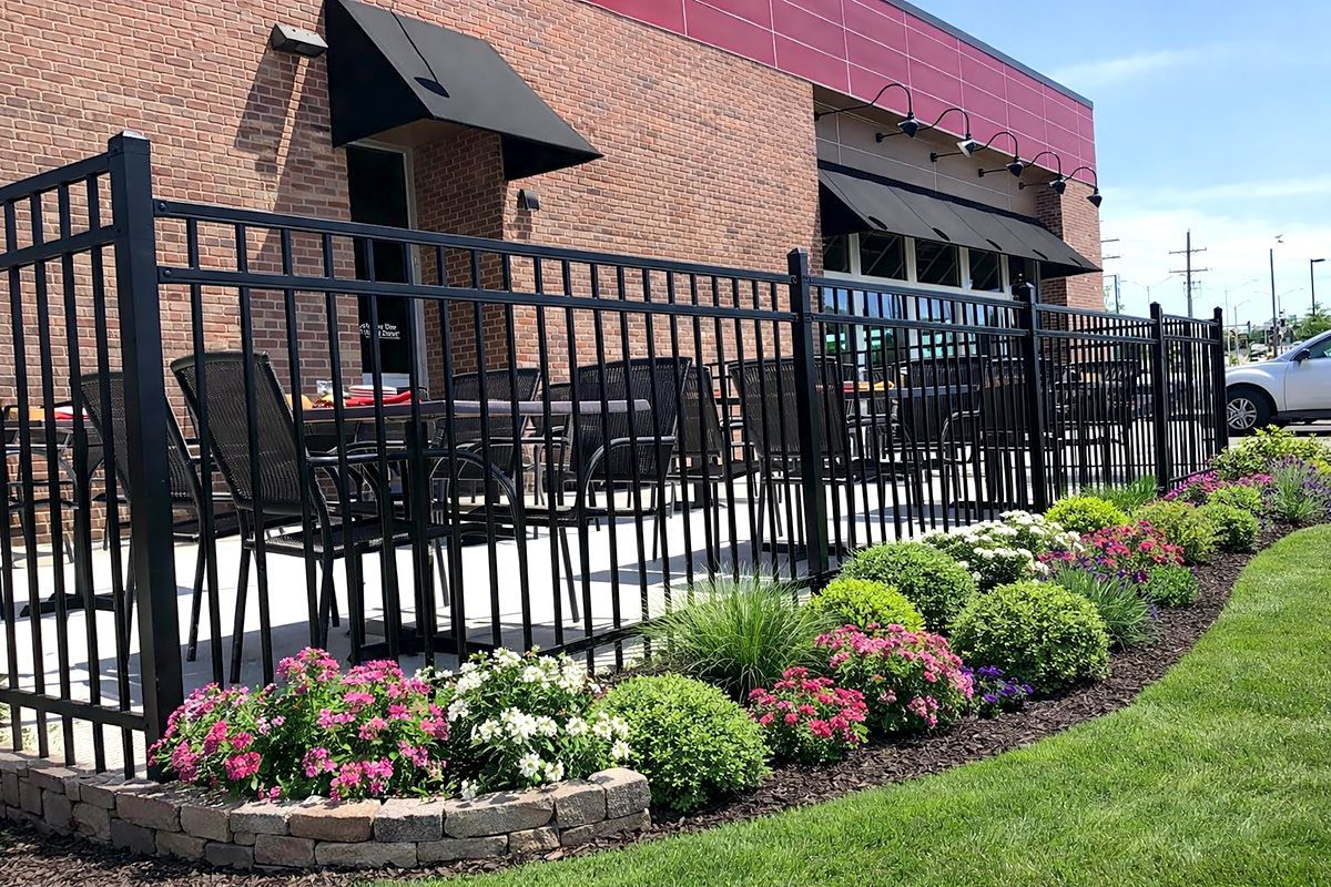 Black fence encloses outdoor dining area with tables, chairs, and flower bed. Building has awnings.