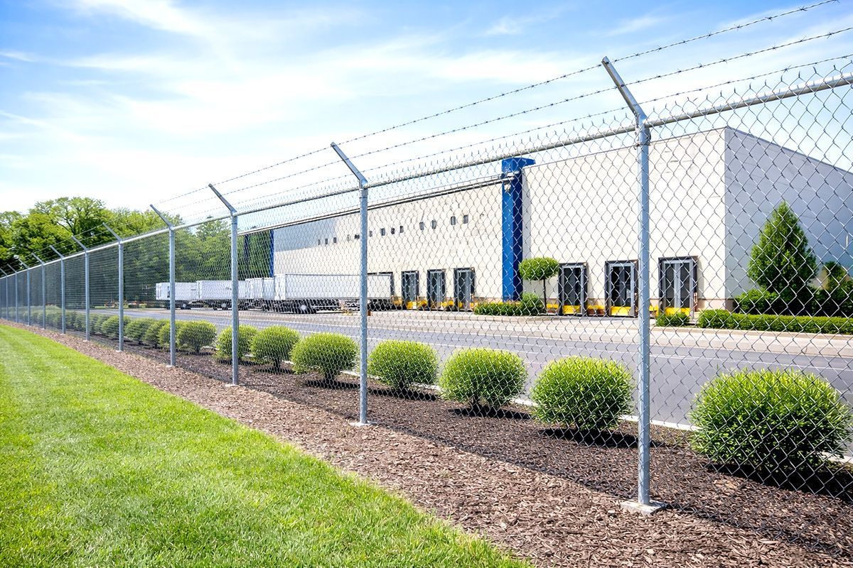 Chain-link fence with barbed wire around a warehouse with loading docks. Green grass and bushes in front.