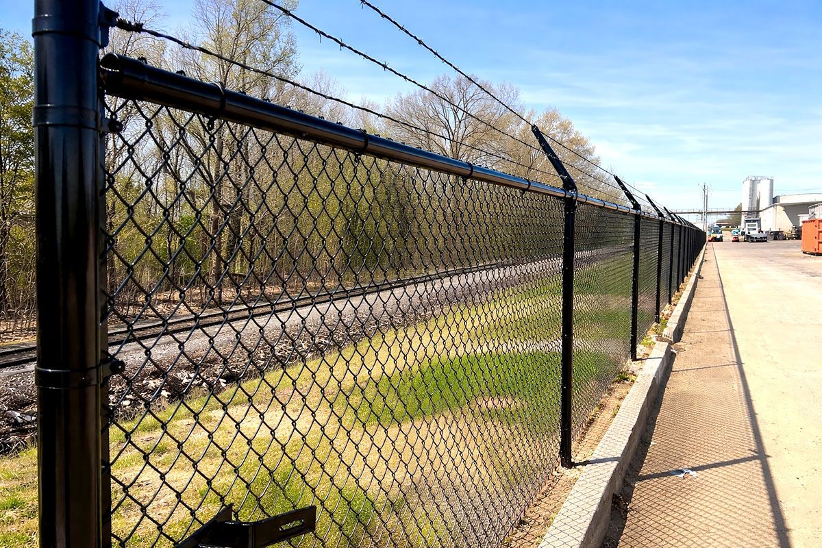 Black chain-link fence with barbed wire on top, bordering a grassy area and railroad tracks.