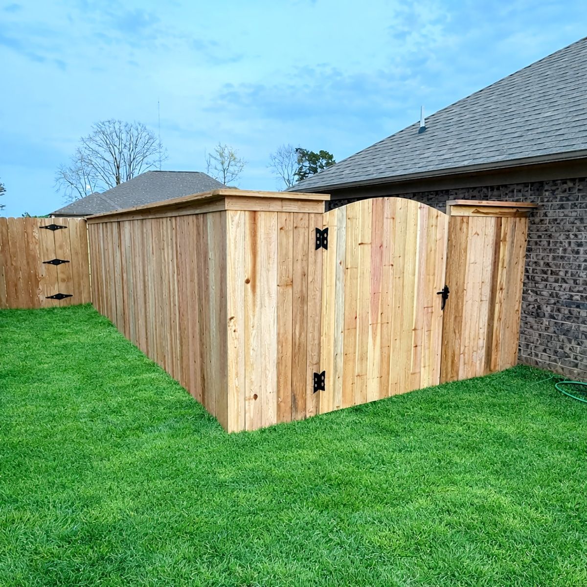 Wooden fence with gate next to a house with green lawn and blue sky.