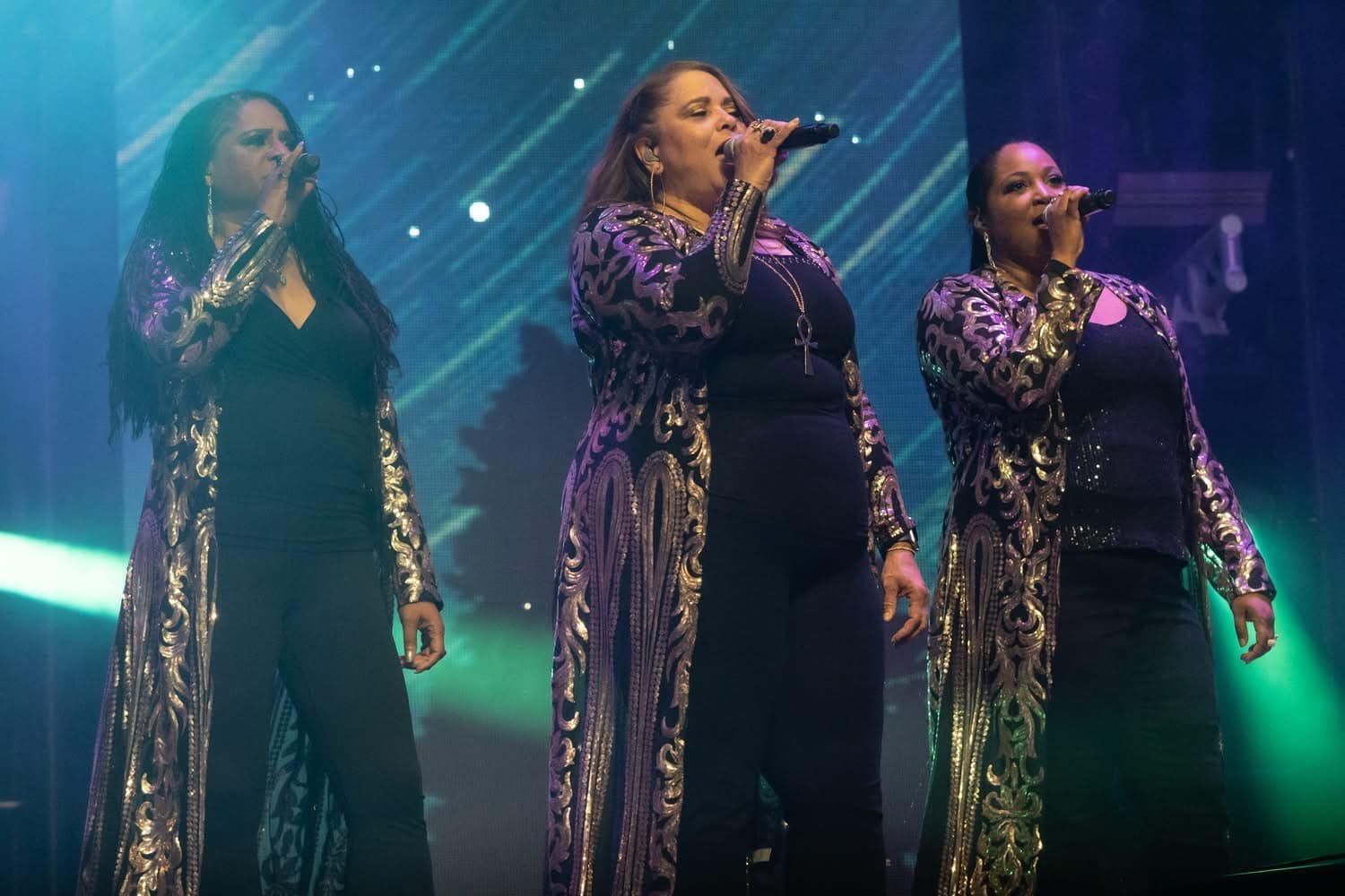 Three women singing on stage; wearing black outfits with sequined jackets.