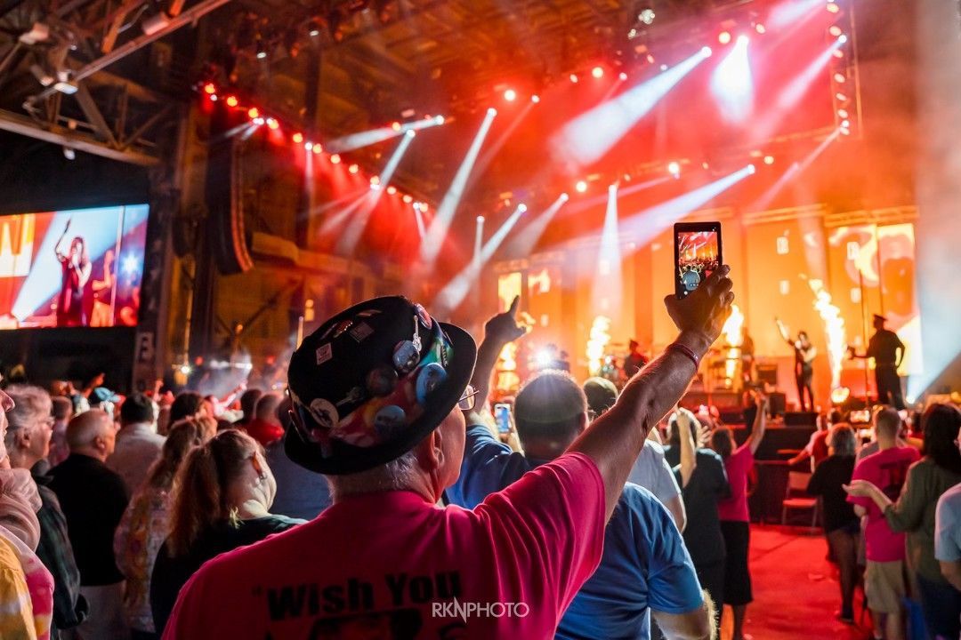 Audience member holding up phone, taking photo of band on stage at concert, red and white stage lights.