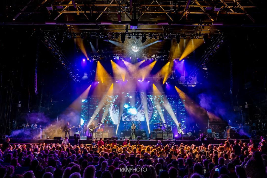Concert stage lit with red and white spotlights shining towards a cheering crowd.