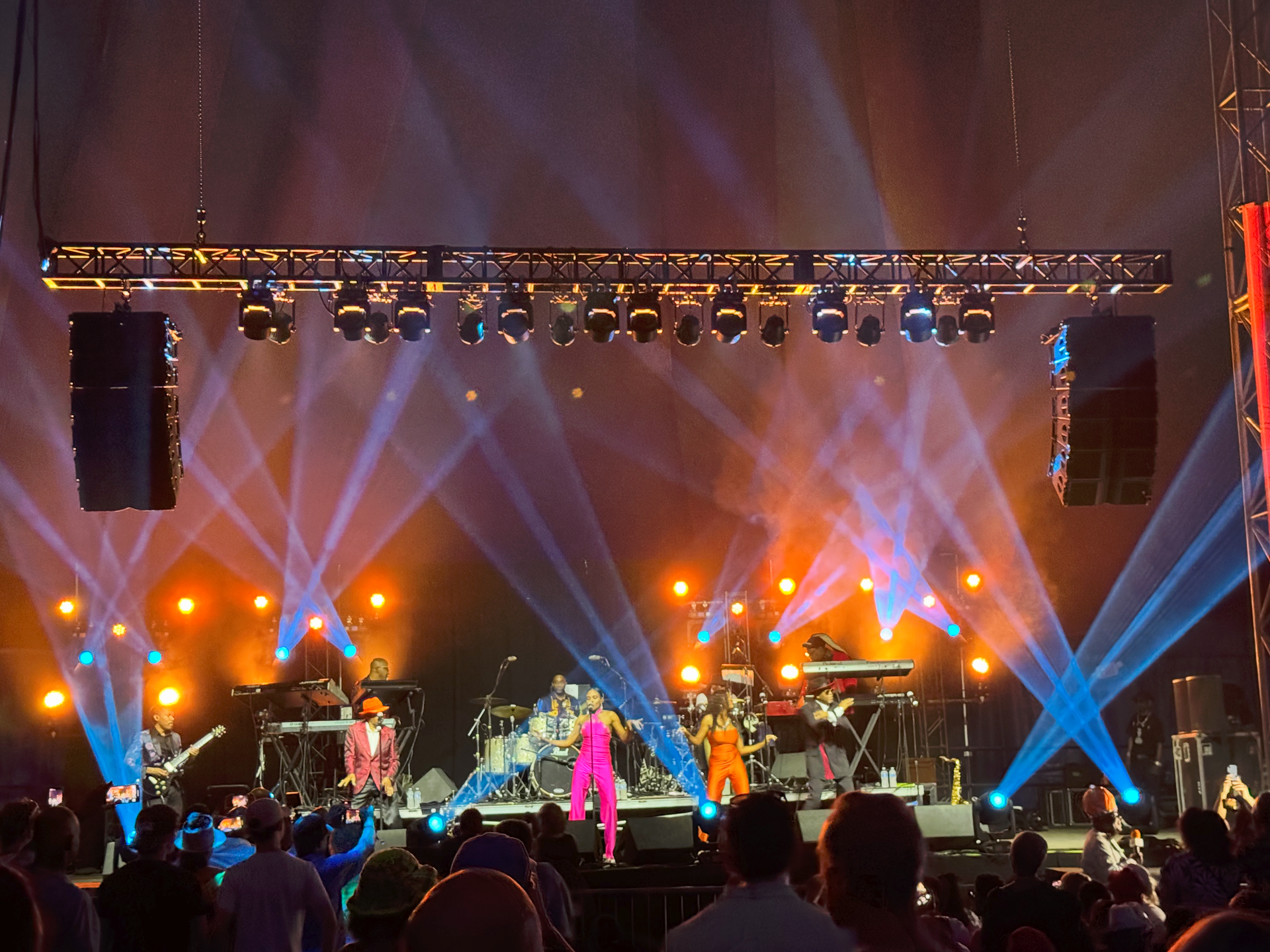 Crowd at a music festival, colorful lights shining, stage with structures, people dancing.