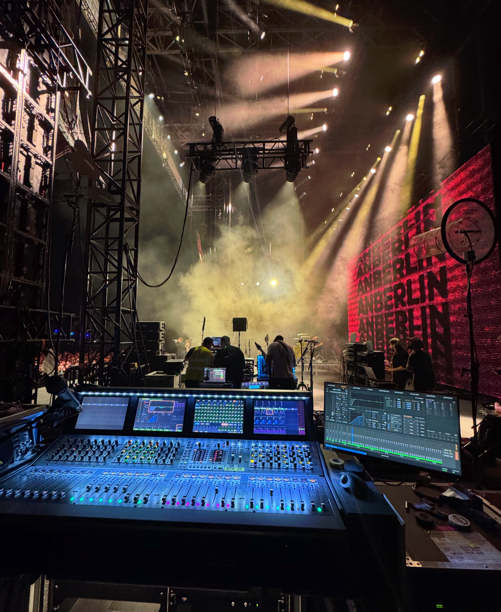 Concert hall scene with hanging speaker stacks. Blue and yellow lights illuminate the crowd. People below.