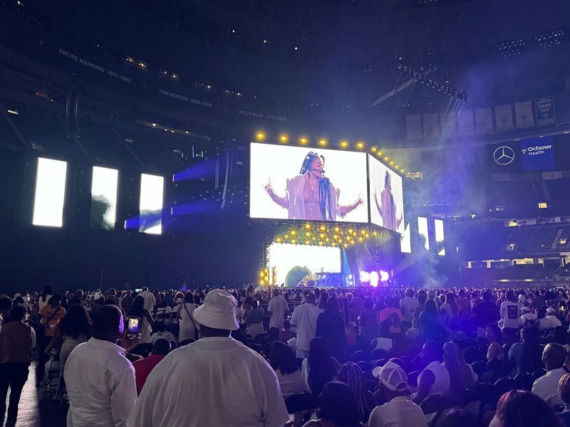 Concert crowd watches a performer on a large screen; stadium setting, stage lights, and people in white clothing.