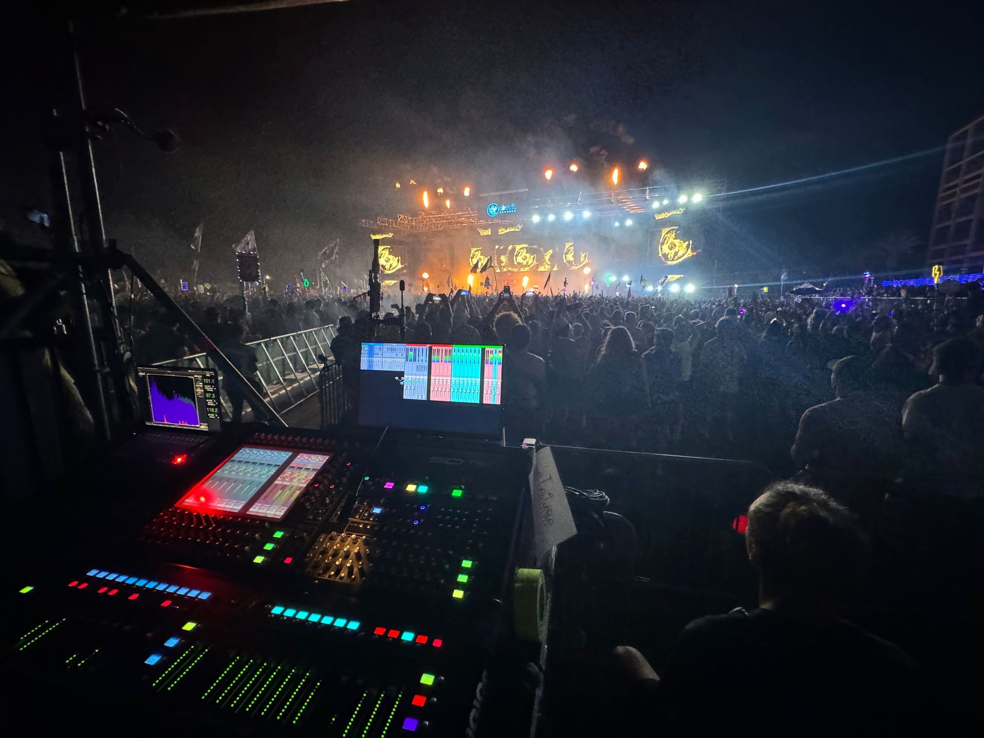 Sound engineer at a large outdoor concert, operating a mixing console with the stage and crowd in the background.
