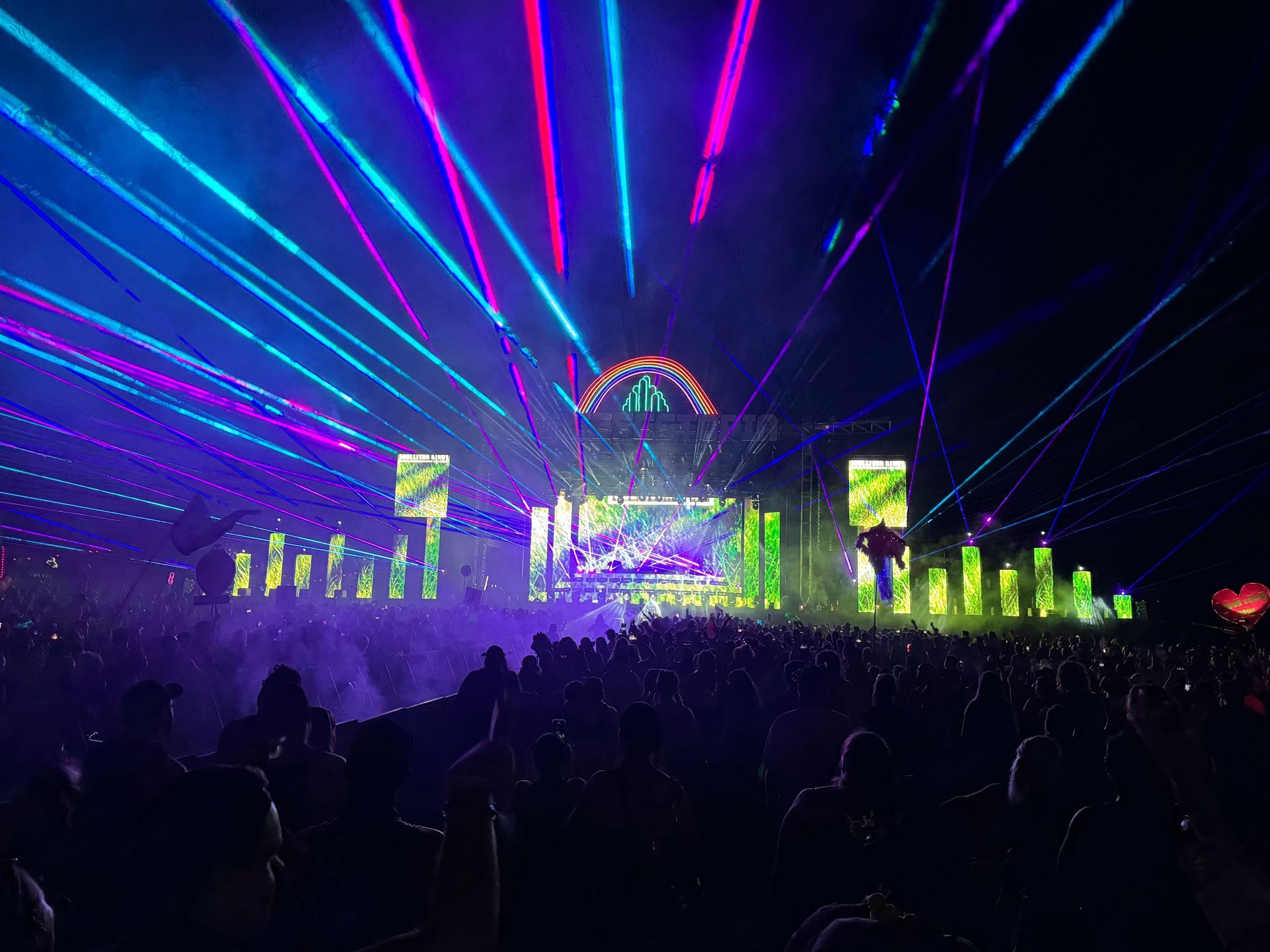 A crowd at a music festival, illuminated by blue and purple laser lights, with a stage in the background.