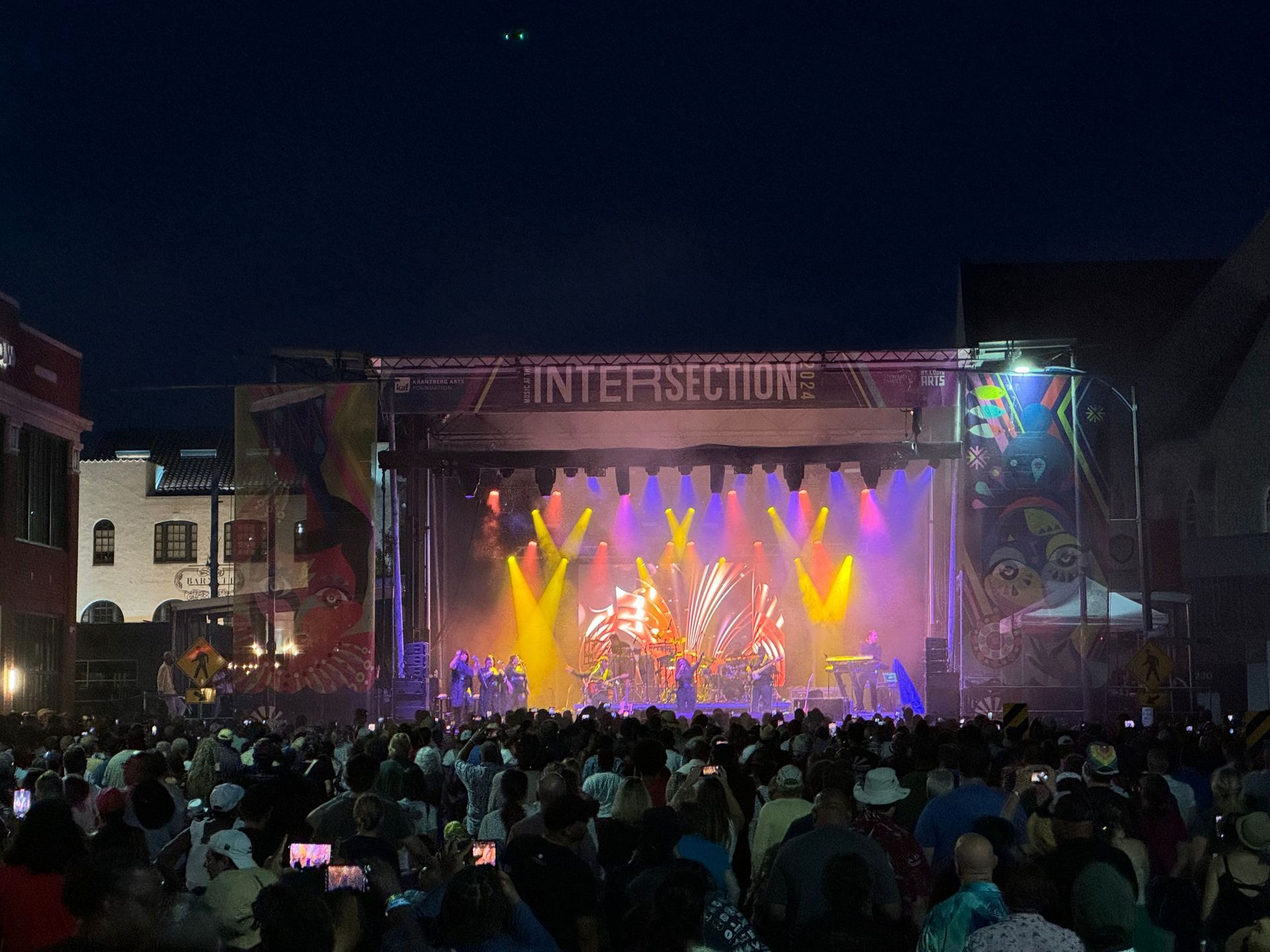 Concert at night, stage with colorful lights, large crowd watching, buildings in background.