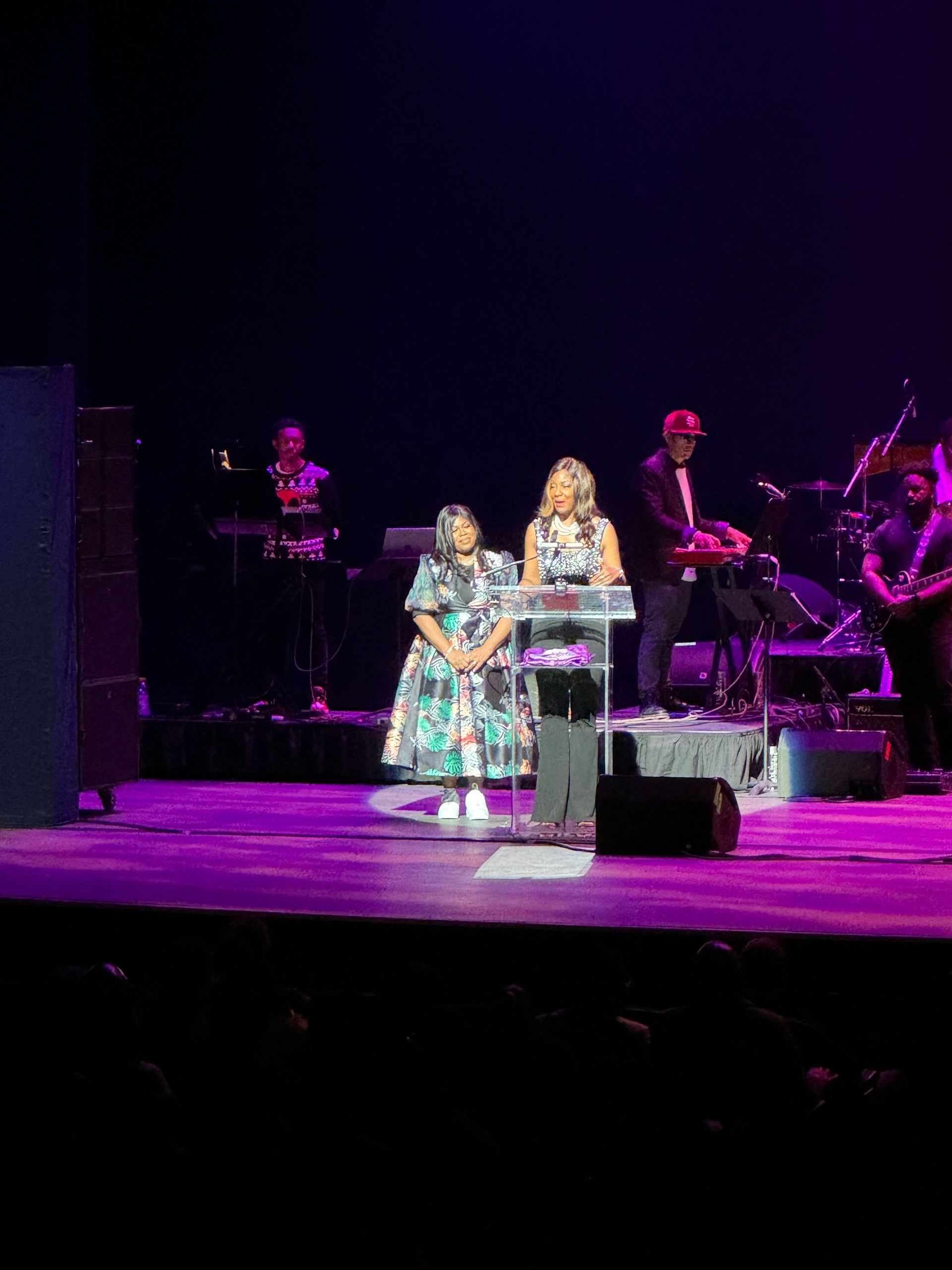 Two women stand at a podium on stage, speaking. A band is visible in the background. Purple stage lighting.