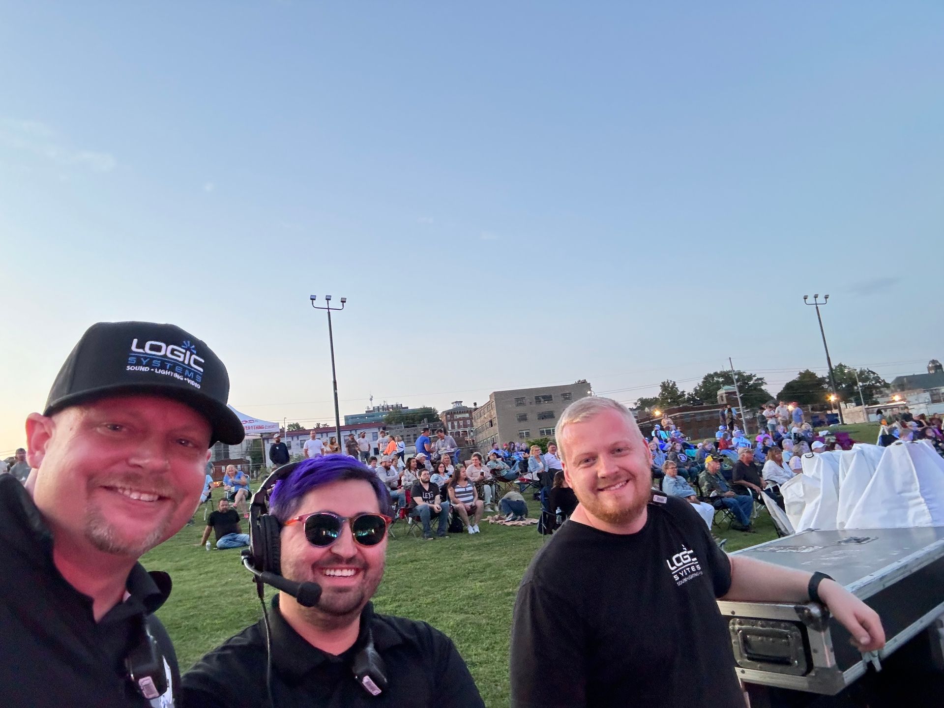 Three men smiling at a large outdoor event, one wearing a headset, under a blue sky.