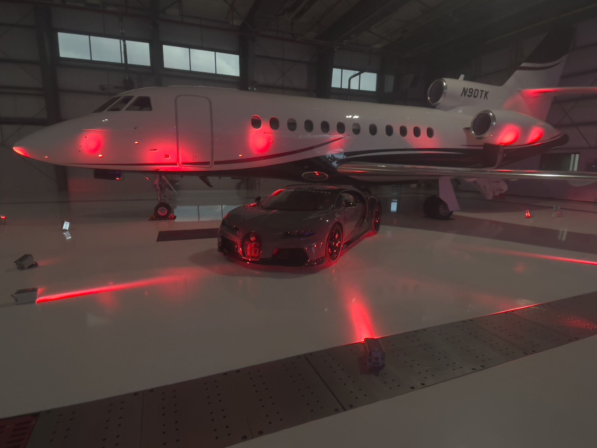A Bugatti sports car parked in front of a private jet, both illuminated with red lights, inside a hangar.