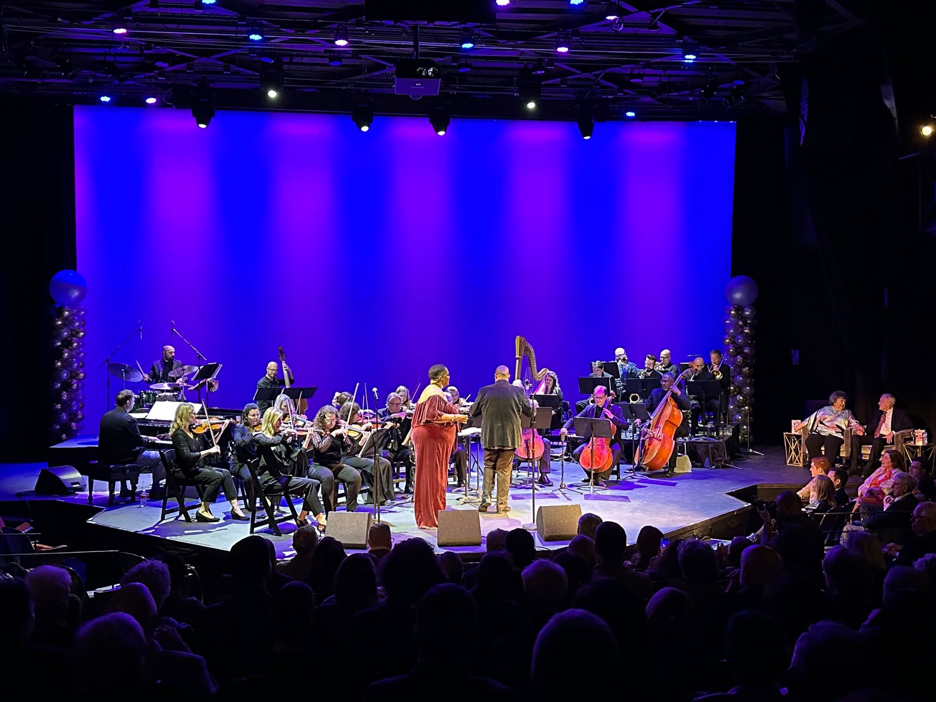 Orchestra performing on stage with conductor, singer, and audience. Purple backdrop.
