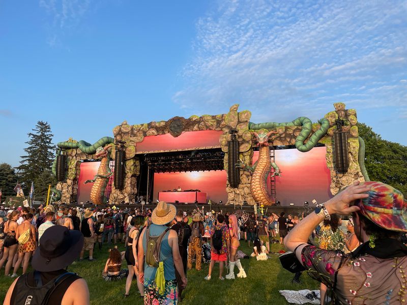 Festival stage with large screen, crowd of people on a grassy field, sunny day.