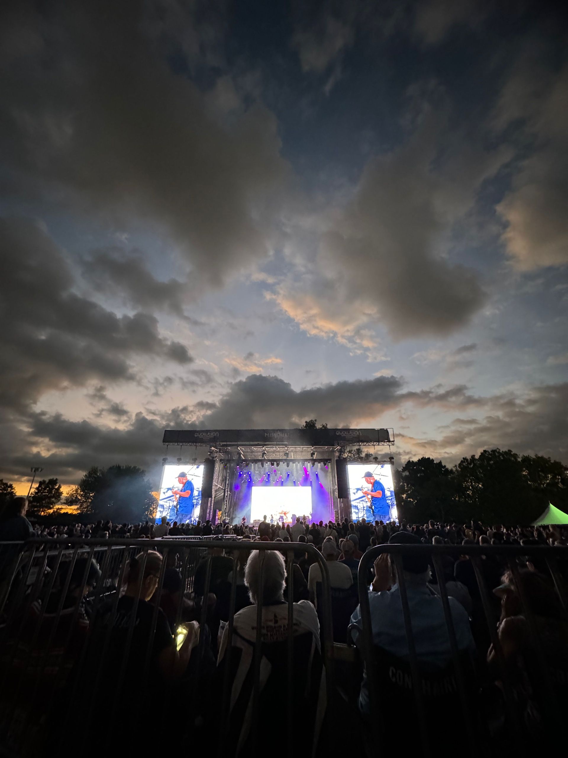 Concert crowd watching a band on stage at dusk. Dark sky with clouds, bright stage lights.