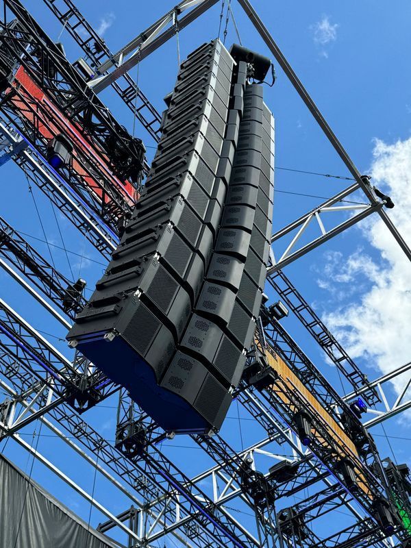 A large speaker array suspended from metal rigging against a blue sky with scattered clouds.