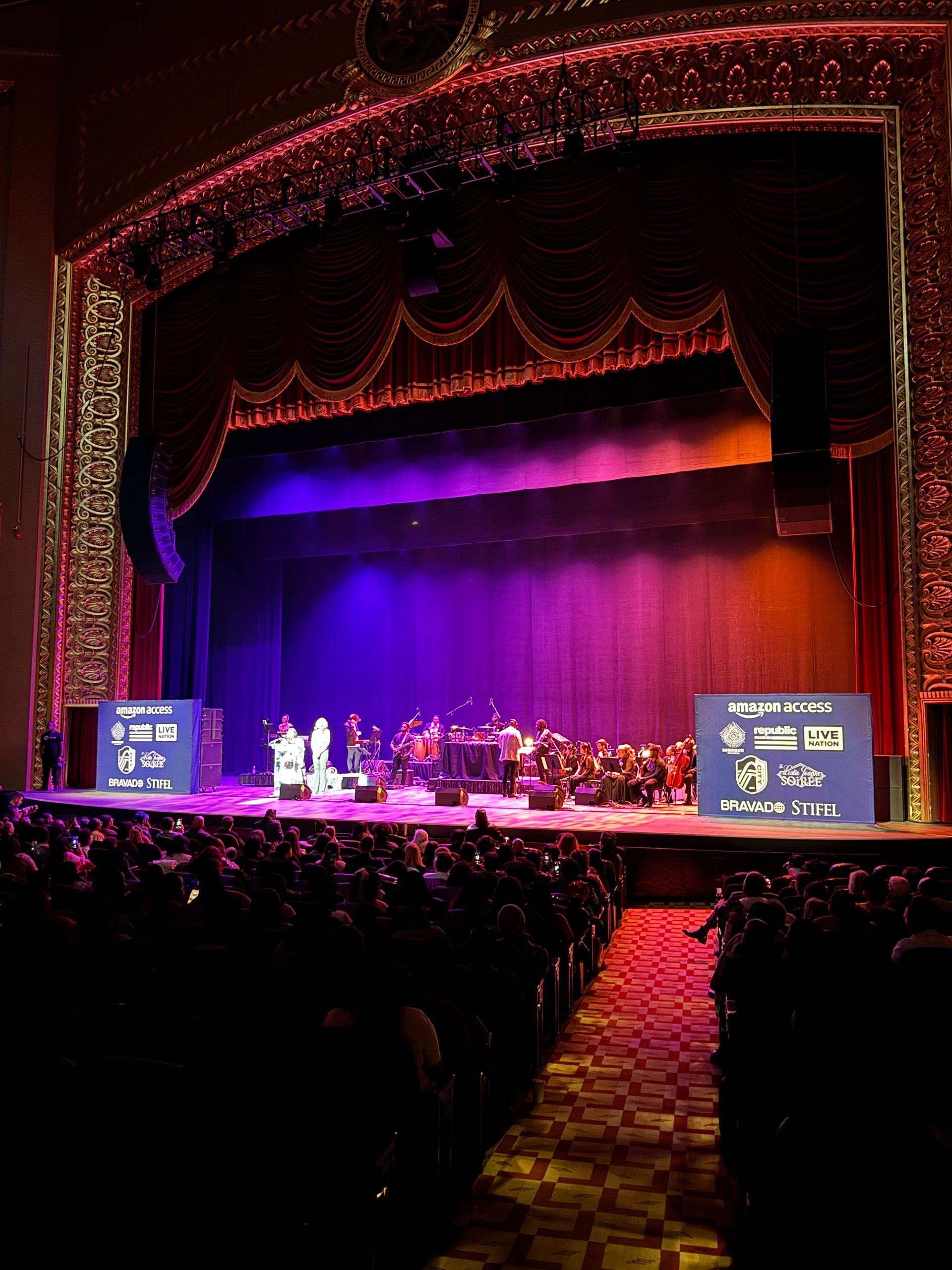 Stage with orchestra, performers, and audience at a formal event, purple/orange lighting.