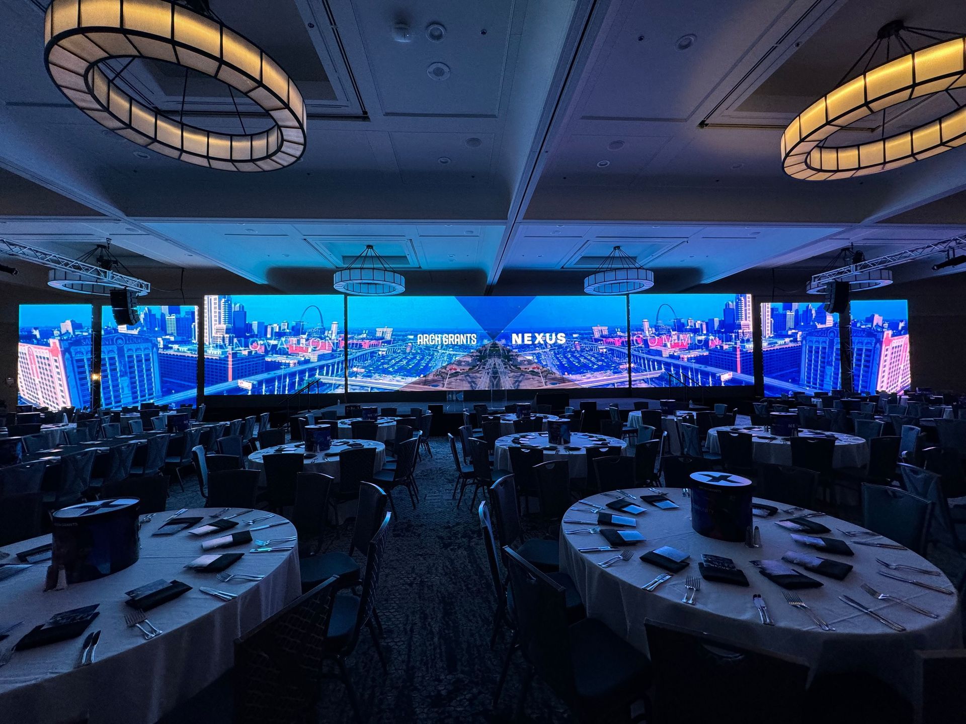 Event hall with large screens displaying a cityscape, round tables set for a meal, and decorative lighting.