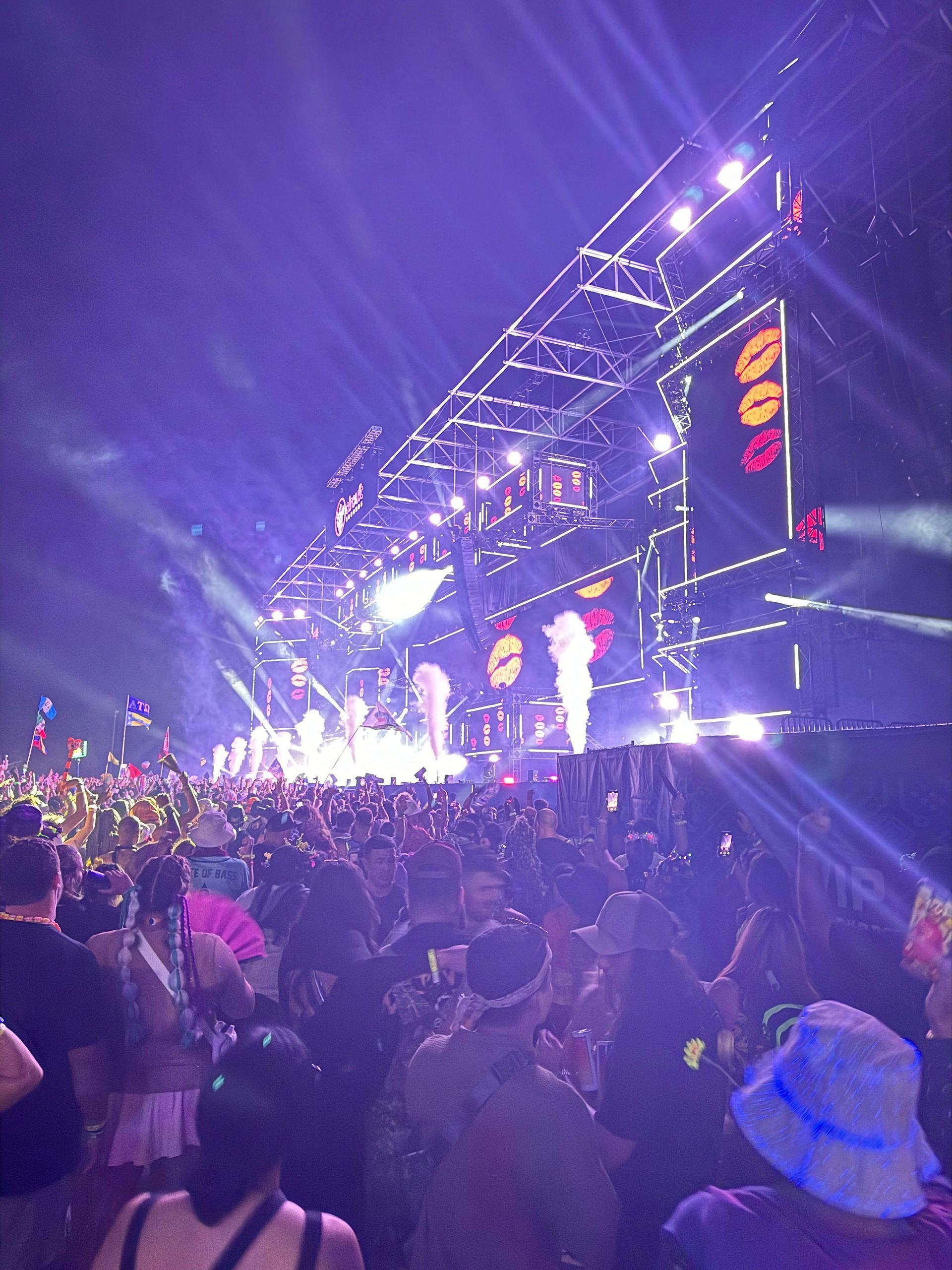 Concert stage with bright lights, smoke, and a large crowd of people dancing.