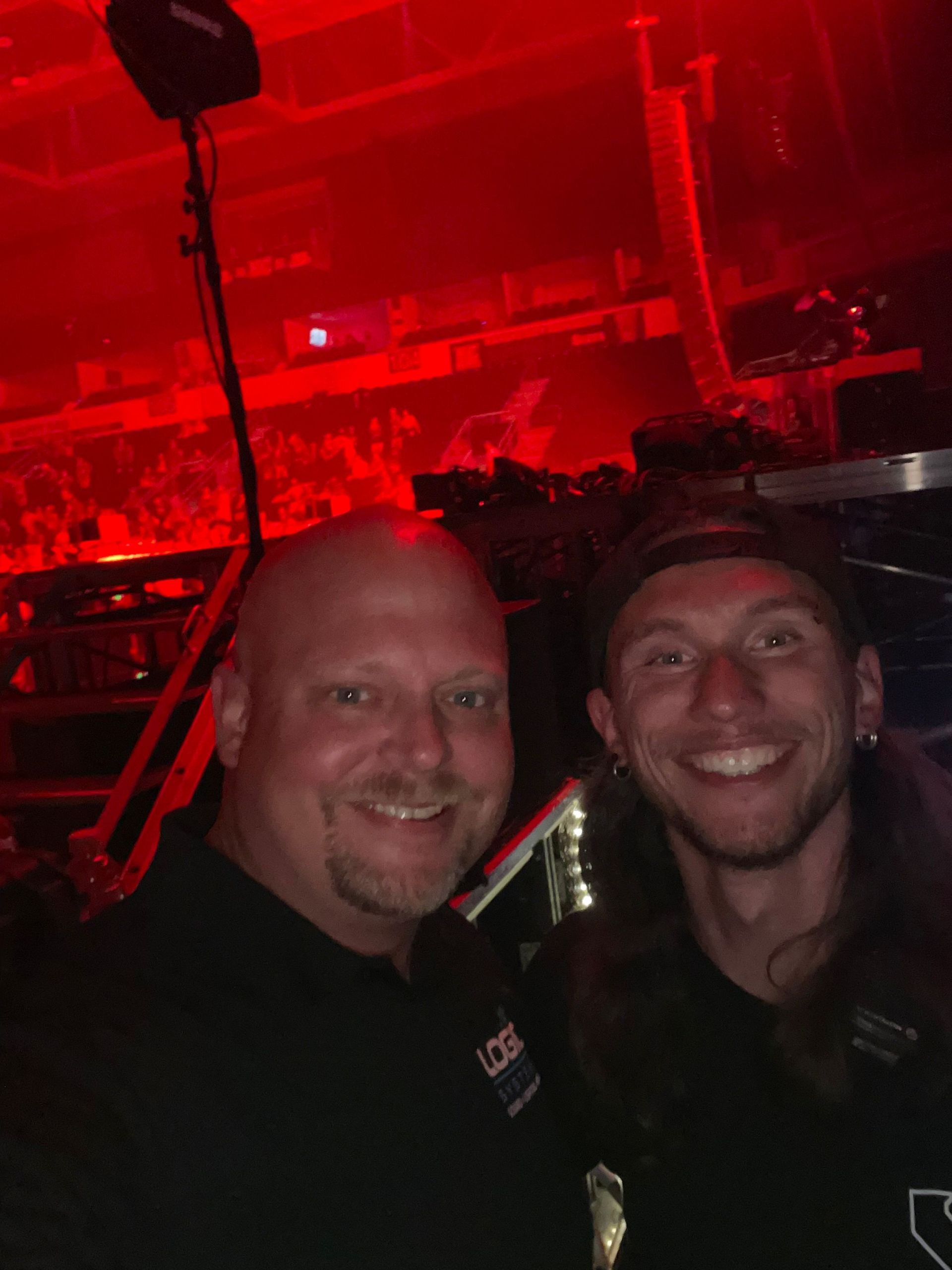 Two men smiling for a selfie at a concert, in red lighting. One bald, the other with long hair.