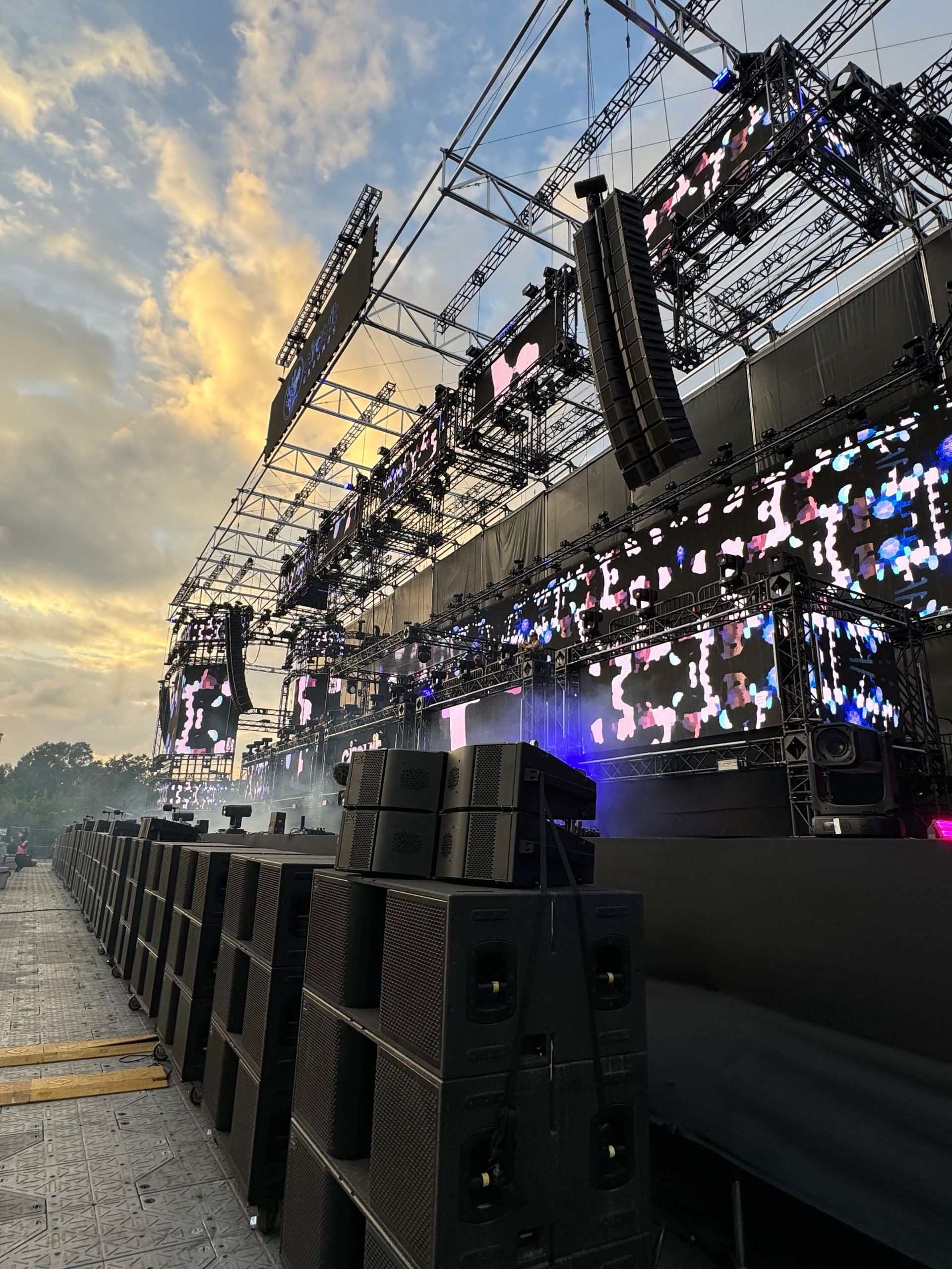 Concert stage set up with many speakers, screens, and metal scaffolding against a cloudy sky.