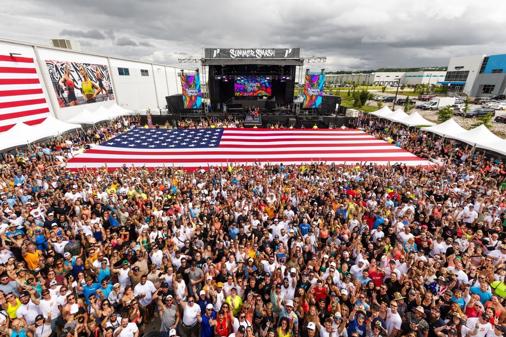 Large outdoor concert with crowd and giant American flag. Stage with band, white tents, sunny sky.