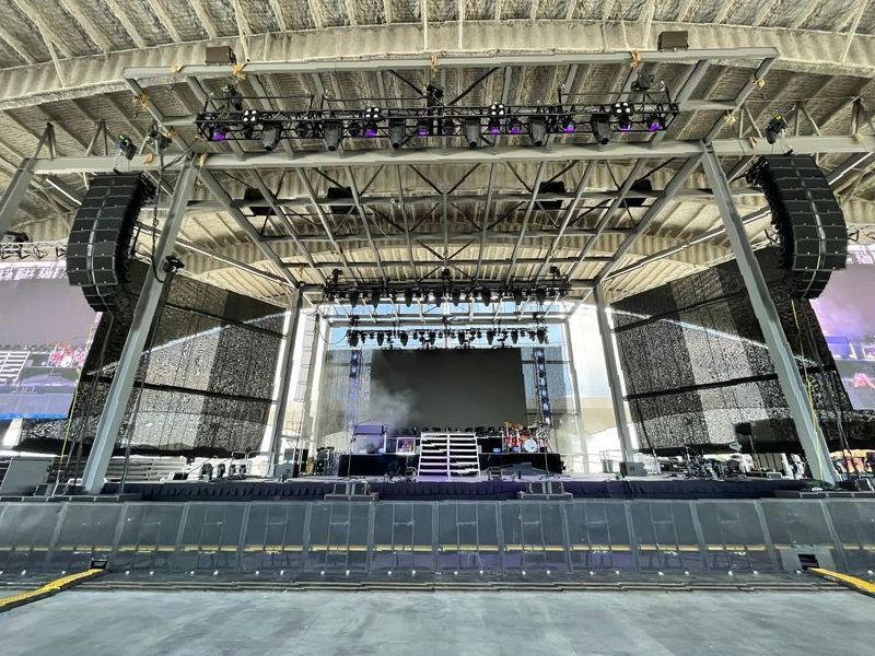 Stage set up inside a stadium; gray structure, speakers, lights, and a screen.