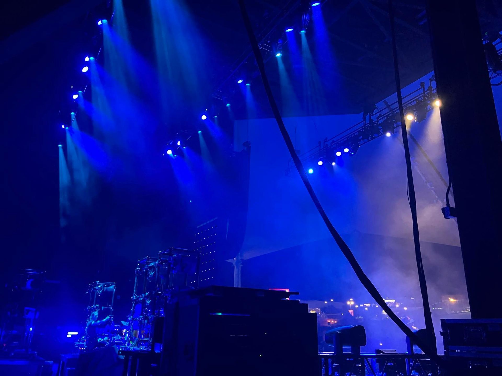 Concert stage with blue spotlights and hazy smoke. A drum set is visible, and a large screen is in the background.
