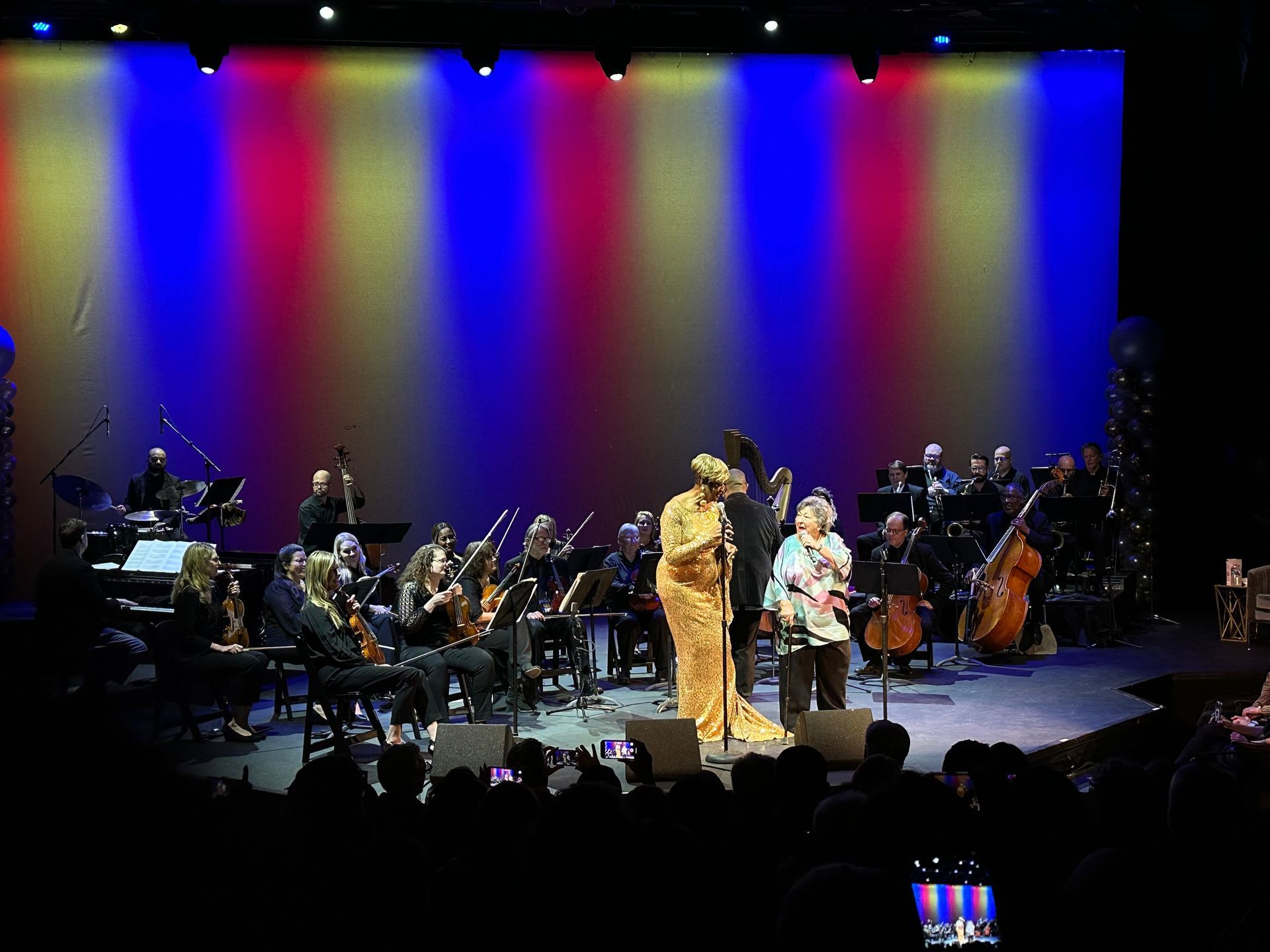 A live musical performance on stage; singer in gold dress with orchestra and crowd below, red, white, and blue lighting.
